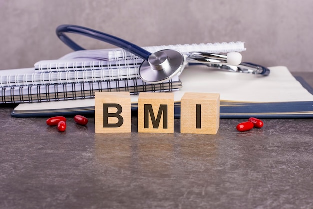 Das Wort BMI steht auf Holzwürfeln in der Nähe eines Stethoskops auf Papierhintergrund. medizinisches konzeptionelles wort, das aus holzelementen mit den buchstaben gesammelt wurde. BMI – Abkürzung für Body-Mass-Index