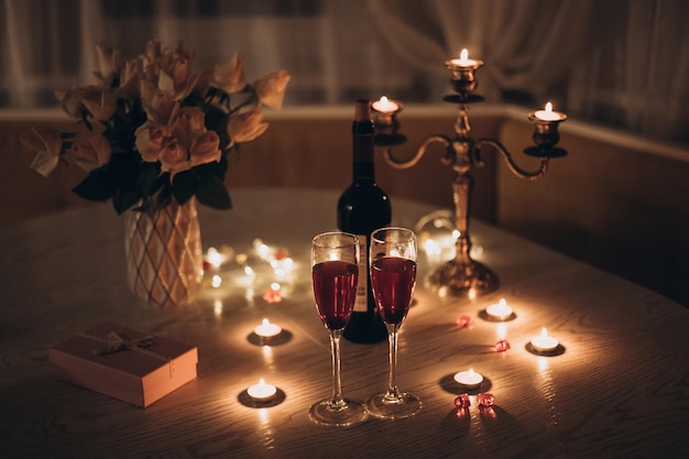 Data à luz de velas. Óculos com buquê de velas de vinho de rosa e caixa de presente na mesa. Jantar romântico à luz de velas em casa à noite