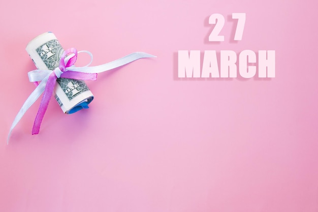 Data do calendário em fundo rosa com notas de dólar enroladas fixadas por fita rosa e azul 27 de março