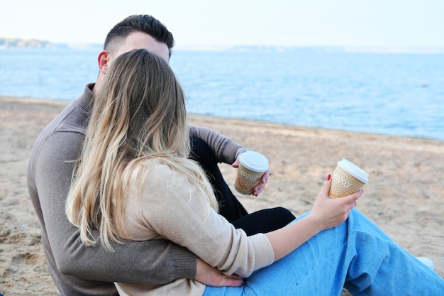 Date am Strand Ein junges Paar in Pullovern sitzt auf einem Plaid, hält Kaffee in Stapeln in den Händen und küsst sich