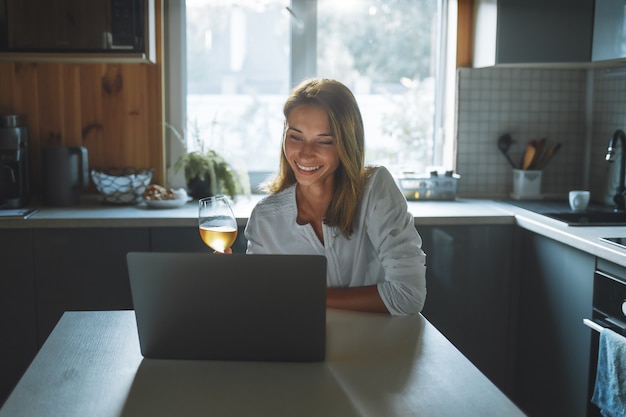 Dating Online-Konzept. Glückliche junge Frau mit Glaswein, der durch soziales Netzwerk spricht, das zu Hause in der Küche sitzt