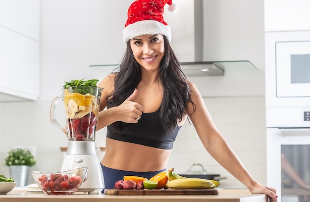 Daumen hoch von einer Frau mit Weihnachtsmütze, die in Trainingskleidung neben einem gesunden Smoothie-Maker voller gesunder Sachen steht.