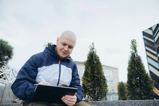 De baixo do homem hipster usando tablet moderno com caneta enquanto cria um novo projeto e sentado na rua