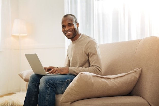 De bom humor. Homem afro-americano de olhos escuros, sorrindo e trabalhando no computador enquanto está sentado no sofá e com uma lâmpada e um tapete ao fundo