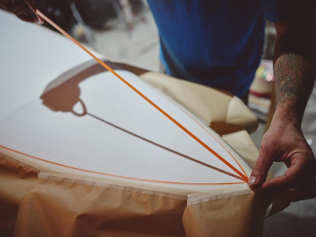 De cima da colheita, um mestre masculino anônimo coloca um elemento decorativo na superfície branca da prancha de surf feita à mão enquanto faz o design da prancheta na oficina.