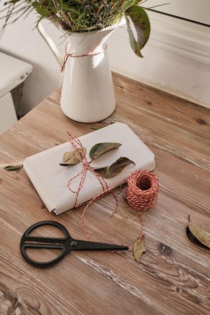 De cima de caixa de presentes feita à mão embrulhada em papel artesanal e decorada com folhas e corda vermelha em uma mesa de madeira