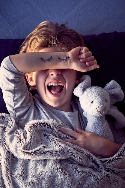 De cima de um menino irritado, deitado na cama com um brinquedo de pelúcia e gritando com a boca aberta enquanto se recusava a dormir e cobria os olhos com a mão com cílios pintados