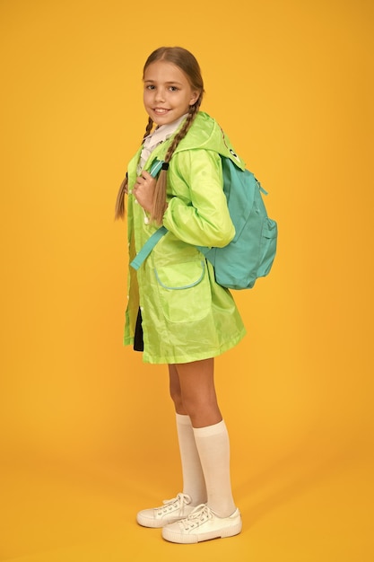 De costas e pronta para ir para a escola Garotinha usa capa de chuva fofa em fundo amarelo Garoto adorável com mochila escolar em dia chuvoso Criança pequena de volta à escola no outono