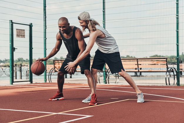 Dê-me a bola! Dois jovens em roupas esportivas jogando basquete e sorrindo enquanto passam o tempo ao ar livre