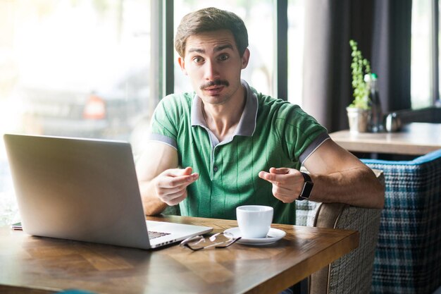 Dê-me dinheiro. Jovem empresário de camiseta verde sentado e olhando para a câmera com gesto de dinheiro e quer seu salário. conceito de negócios e freelancer. tiro interno perto da grande janela durante o dia.