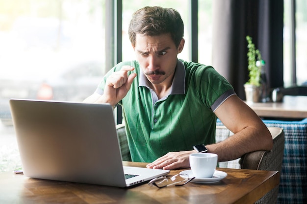 Dê-me mais um pouco de jovem empresário de camiseta verde sentado e olhando para a tela do laptop na chamada de vídeo e pedindo para lhe dar mais tempo tiro interno do conceito de negócios perto da grande janela durante o dia