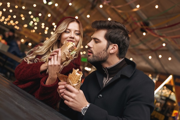 Dê-me uma mordida. Jovem casal está comendo sanduíches no mercado de comida de rua. Estação fria. Foto de close-up de um casal comendo em um restaurante