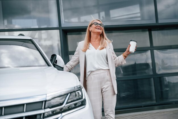 De pé ao ar livre contra a construção e segurando a bebida A mulher com roupas formais brancas está na concessionária de carros