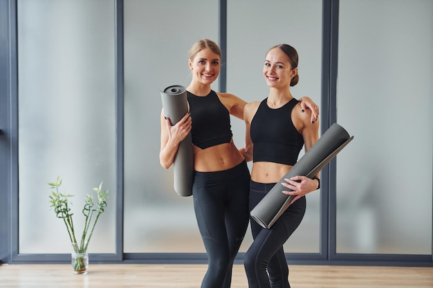 De pé com tapetes nas mãos Duas mulheres em roupas esportivas e com corpos esbeltos têm dia de ioga fitness dentro de casa juntos