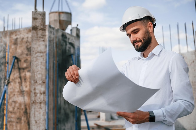 Foto de pé e segurando o plano nas mãos o homem de uniforme está trabalhando no canteiro de obras