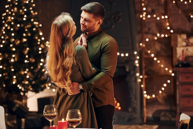De pé na sala decorada de Natal Jovem casal adorável tem jantar romântico dentro de casa juntos
