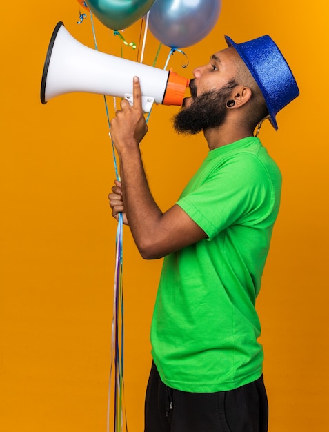 De pé na vista de perfil, um jovem afro-americano com chapéu de festa segurando balões fala no alto-falante