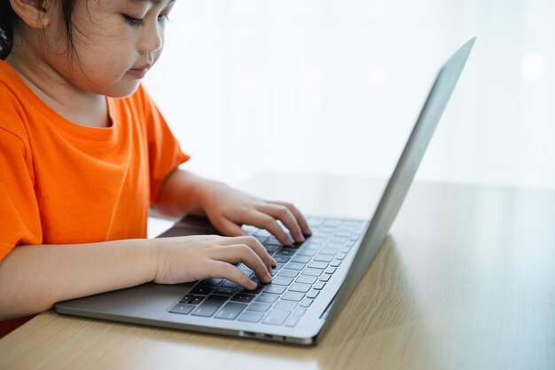 De perto, uma menina asiática vestindo uma camiseta laranja usa um laptop e estuda e-learning on-line em uma mesa de madeira na sala de estar em casa.