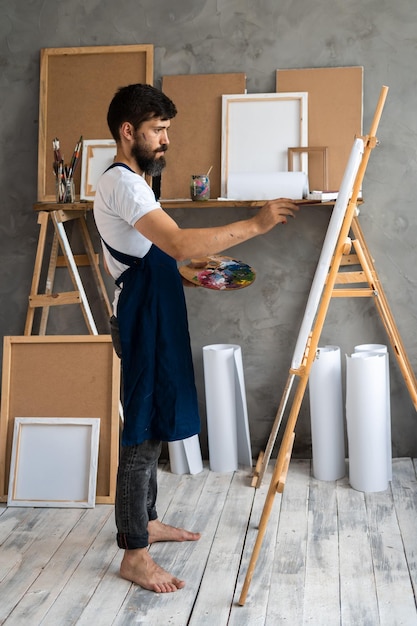 Foto de um artista masculino barbudo no estúdio, descalço e de avental, fica em um cavalete sobre uma tela com um pincel e uma paleta com tintas e está ocupado com seu amado hobi