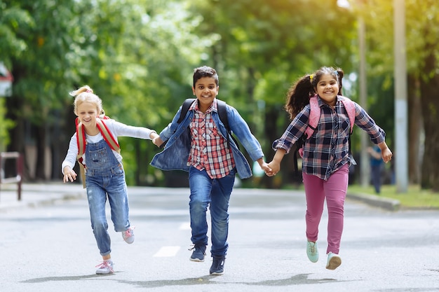 De volta à escola. Grupo racial misturado de estudantes felizes da escola primária com as trouxas que correm guardando as mãos fora. Conceito de educação primária.