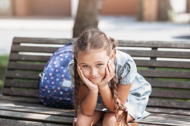 De volta à escola uma colegial bonitinha em um vestido com tranças e mochila azul grande está sentado em um banco no pátio da escola uma garotinha está indo para a primeira série