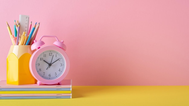 De volta ao conceito de escola Foto de material escolar no despertador de mesa amarelo suporte para régua de lápis de canetas e pilha de cadernos no fundo da parede rosa com espaço vazio