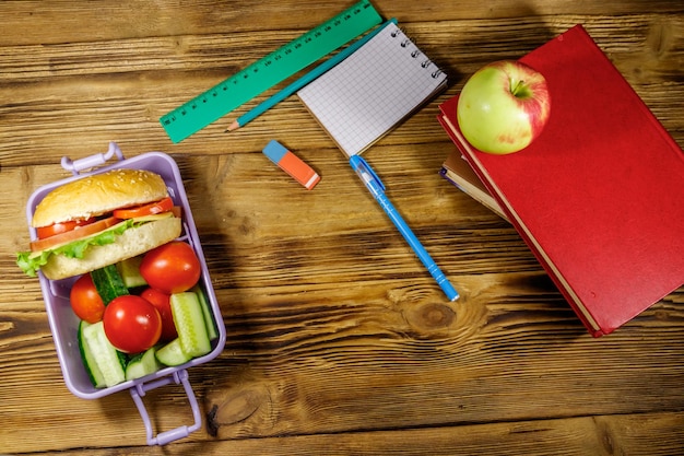 Foto de volta ao conceito de escola material escolar livros maçã e lancheira com hambúrgueres e legumes frescos em uma mesa de madeira vista superior