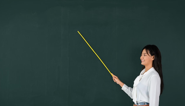 De volta ao conceito de escola. Professora asiática sorrindo com bastão de madeira apontando para o quadro-negro na escola em sala de aula, feliz e bela jovem segurando o ponteiro para o painel traseiro