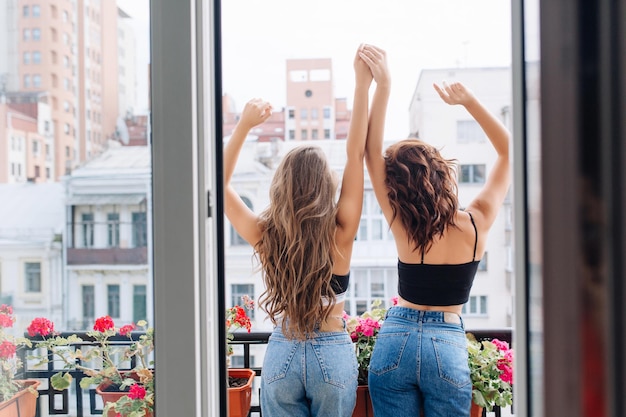 De volta, duas mulheres elegantes em jeans e camiseta preta na varanda. Garotas atraentes com corpos esguios, segurando as mãos, se divertindo no cityview. Relaxamento, amigos, emoções brilhantes