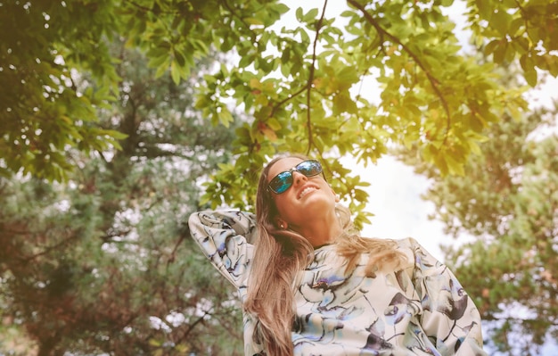 Foto debajo de la vista de la joven y bella mujer rubia con gafas de sol tocando su pelo largo sobre el cielo y los árboles. libertad y disfrute del concepto.