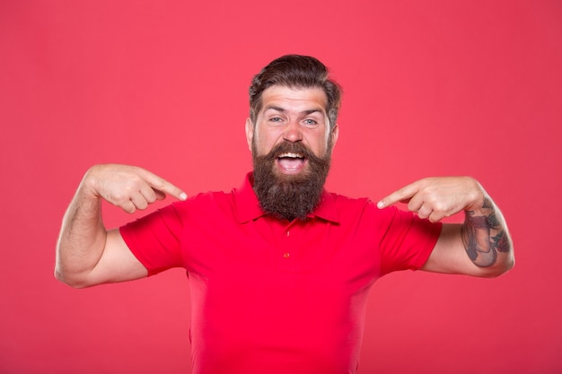 Debe tener. Hipster feliz apuntando al fondo rojo de la camiseta. Señalar con los dedos índices. Apuntando a la publicidad. Gesto señalador. Márketing y promoción. Tienda de ropa masculina. Tienda y compras, copie el espacio.