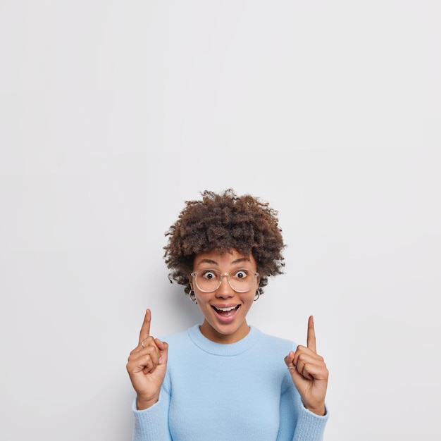 Deberías echarle un vistazo a la hermosa mujer de cabello rizado positivo que señala con el dedo índice arriba, elige el producto, se siente asombrado, usa anteojos transparentes y un puente casual azul aislado sobre fondo blanco.