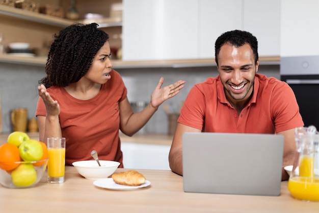 Decepcionada y enojada esposa afroamericana gritando al marido sonriente jugando en la laptop