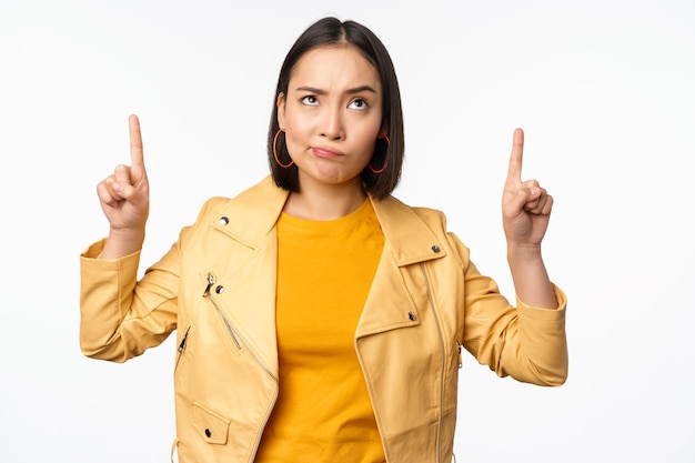 Decepcionada modelo femenina coreana frunciendo el ceño y haciendo una mueca escéptica apuntando hacia arriba y mirando molesta a la pancarta en la parte superior de pie sobre fondo blanco.