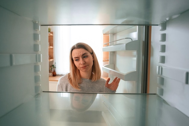 Foto decepcionado por un refrigerador vacío sin comida en él una hermosa joven está en la cocina