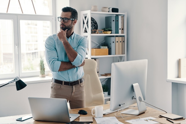 Decisão difícil de tomar. Jovem empresário moderno mantendo a mão no queixo e olhando para longe enquanto trabalha no escritório