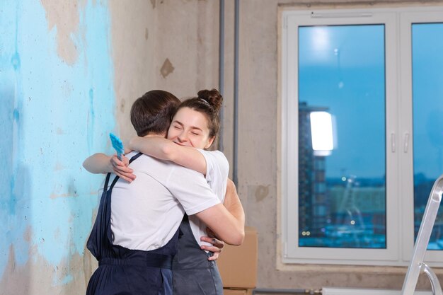 Decoração de casa DIY conceito de renovação Casal feliz durante o reparo em uma sala vazia de uma nova casa pintando a parede juntos Marido e mulher se abraçando na nova casa Família feliz fazendo reforma em casa