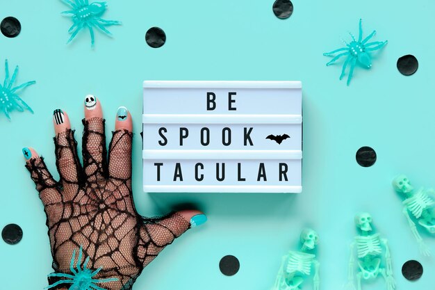 Decoração de Halloween e texto motivador Seja Spooktacular na mesa de luz. Aranha por lado na luva de malha com manicure de Halloween. Postura plana sobre fundo verde menta com esqueletos verdes pálidos e confetes pretos.