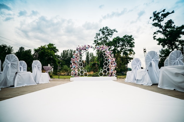 decoración de una ceremonia al aire libre en un jardín verde, un arco de flores frescas, un camino blanco para los recién casados, sillas con fundas de tela blanca