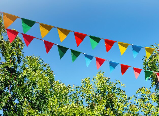 decoración de cuerdas de banderas coloridas en el follaje verde de los árboles en el fondo de la fiesta de verano del cielo azul AI generado