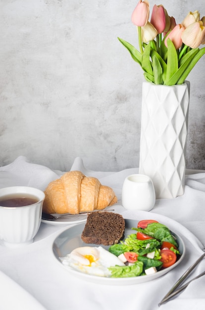 Decoración de mesa de primavera por la mañana. Ensalada en plato, huevo, taza de café y croissant, tulipanes frescos en florero sobre fondo de mantel blanco limpio. Cubierto de mesa de desayuno en color blanco.