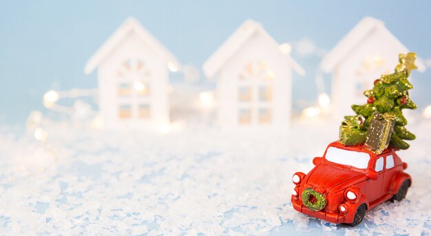 Decoración navideña - coche retro rojo sobre la nieve lleva el árbol de Navidad cerca de las casas