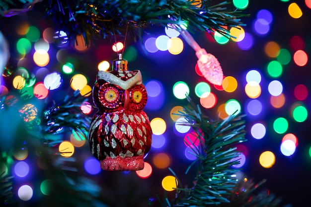 Decoración navideña en forma de un búho rojo colgando de un árbol de Navidad en el fondo, brillan muchas guirnaldas en diferentes colores.