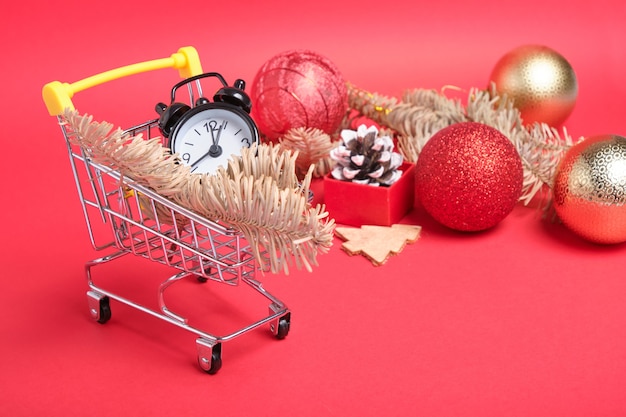 Decoración navideña y pequeño reloj despertador negro en un mini carrito de la compra sobre una superficie roja