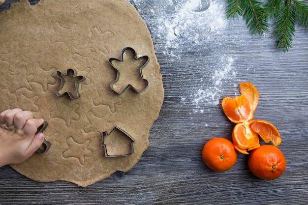 Decorações de Ano Novo e Natal em uma superfície de madeira com tangerinas e uma árvore de Natal As mãos de uma criança pequena fazem moldes para cozinhar pão de gengibre na forma de um homem