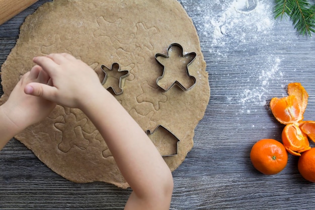 Decorações de Ano Novo e Natal em uma superfície de madeira com tangerinas e uma árvore de Natal As mãos de uma criança pequena fazem moldes para cozinhar pão de gengibre na forma de um homem