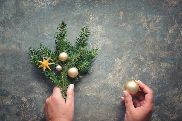 Decorações de Natal planas com as mãos segurando ramos de pinheiro decorados com bugigangas douradas, bugigangas e estrelas. Postura plana na mesa texturizada escura.