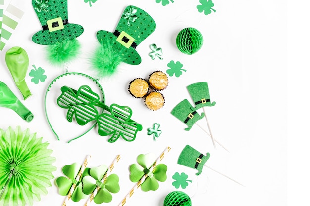 Decorações e adereços para a festa do Dia de São Patrício. Decorações de papel verde e dourado, aro de cabelo com chapéus, folha de trevo, óculos de sol, balões, confetes e doces em fundo branco. Camada plana, vista superior