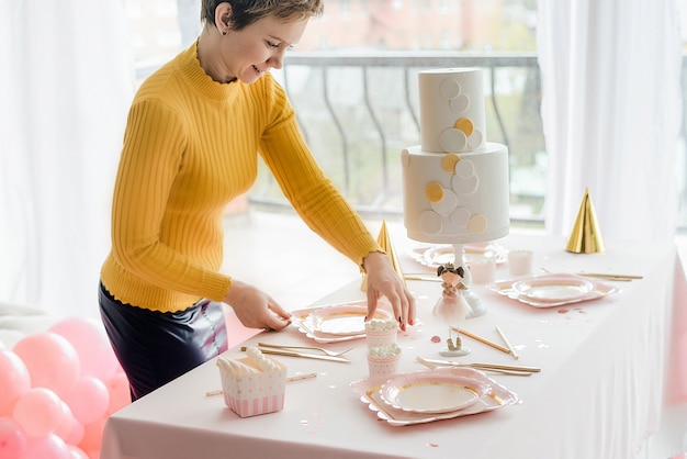 Decorador perto da mesa de festa em tons pastel com toalha rosa, pratos coloridos de papel, copos e talheres de ouro. decoração de festa de aniversário de menina, bonés festivos.