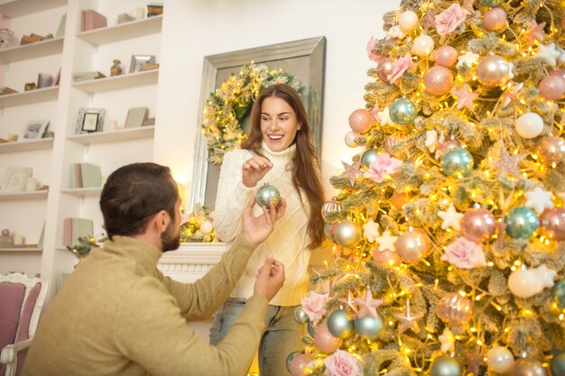 Decorar a árvore de Natal. Jovem casal feliz colocando bolas de Natal em uma árvore de Natal